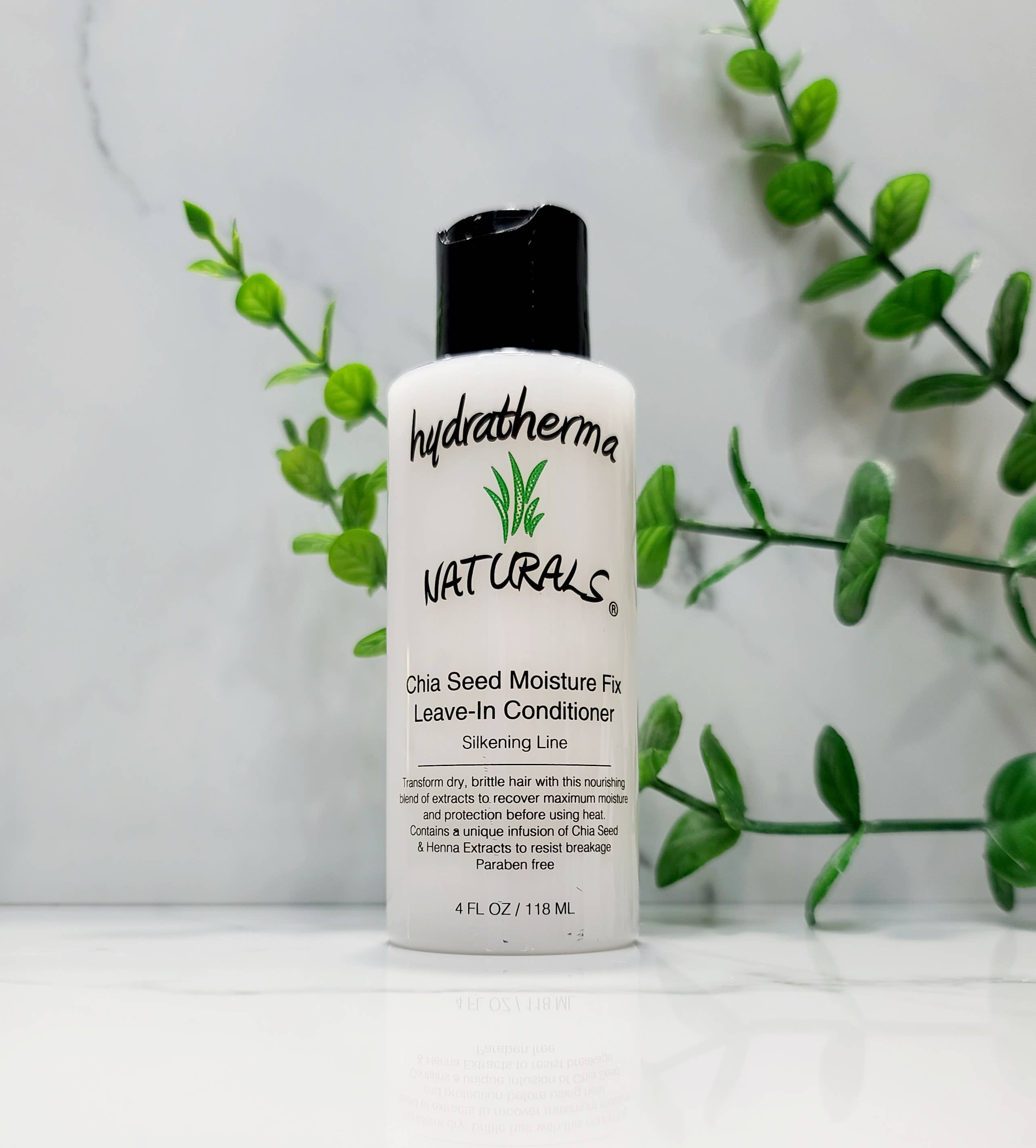 A bottle of Hydratherma Naturals Chia Seed Moisture Fix Leave-In Conditioner stands in front of green sprigs against a white marble backdrop.